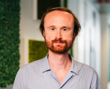 A person with a beard and a slight smile stands indoors, wearing a light blue shirt. The background features blurred green and white panels.