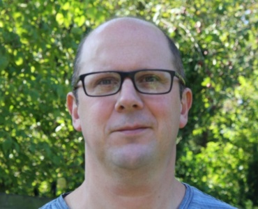 A person wearing glasses and a blue shirt stands in front of vibrant green foliage.