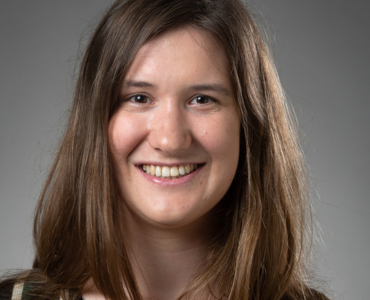 A woman with long brown hair smiles warmly against a gray background.