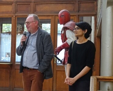 A man speaks into a microphone alongside a smiling woman. Behind them, a figure in a red costume stands. The setting is indoors, with wooden walls.