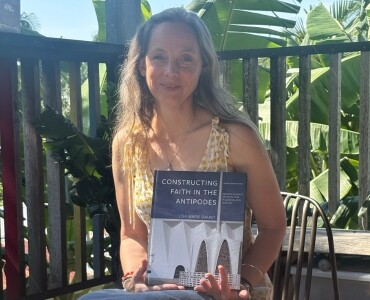 A woman with long hair, wearing a yellow patterned dress, sits outdoors holding a book titled 