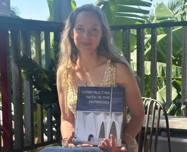 A woman with long hair, wearing a yellow patterned dress, sits outdoors holding a book titled