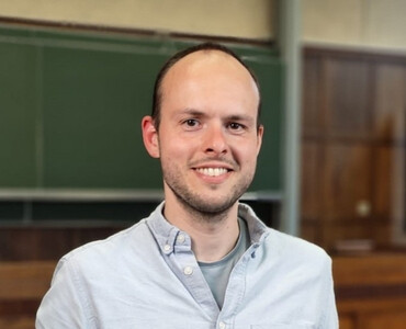 A man with a friendly smile stands in a classroom, wearing a light blue shirt. A chalkboard and wooden paneling are visible in the background.