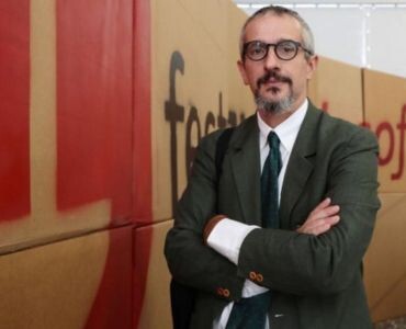A man with glasses and a beard stands with arms crossed, wearing a suit and tie.