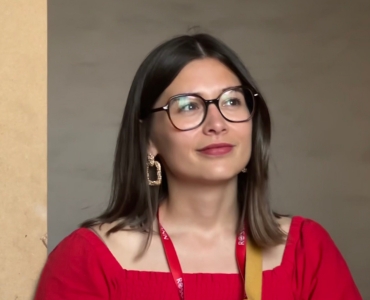 A woman with long brown hair and glasses wearing a red top looks thoughtfully upward.