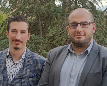 Two men stand outdoors in front of tall, lush trees.