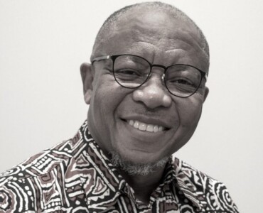 Smiling man with glasses wearing a patterned shirt, set against a plain background.