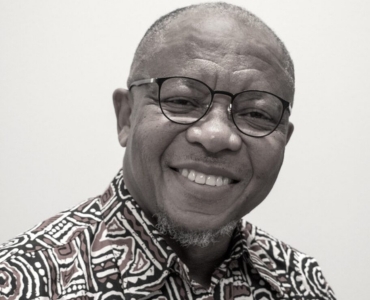 Smiling man with glasses wearing a patterned shirt, set against a plain background.