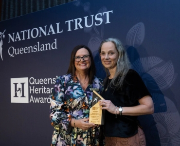 Two women pose together holding an award in front of a backdrop that reads 