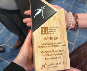 A close-up of a trophy held by two hands. The trophy is made of wood with a black top section, displaying the words "WINNER" and detailing the award for "Heritage Reports, Resources, Publications" awarded to Dr. Lisa Marie Daunt by the National Trust Queensland.