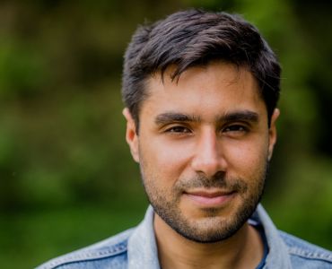 Close-up outdoor headshot of a person facing the camera. The person has short dark hair and light facial hair and is wearing a light blue collared shirt.