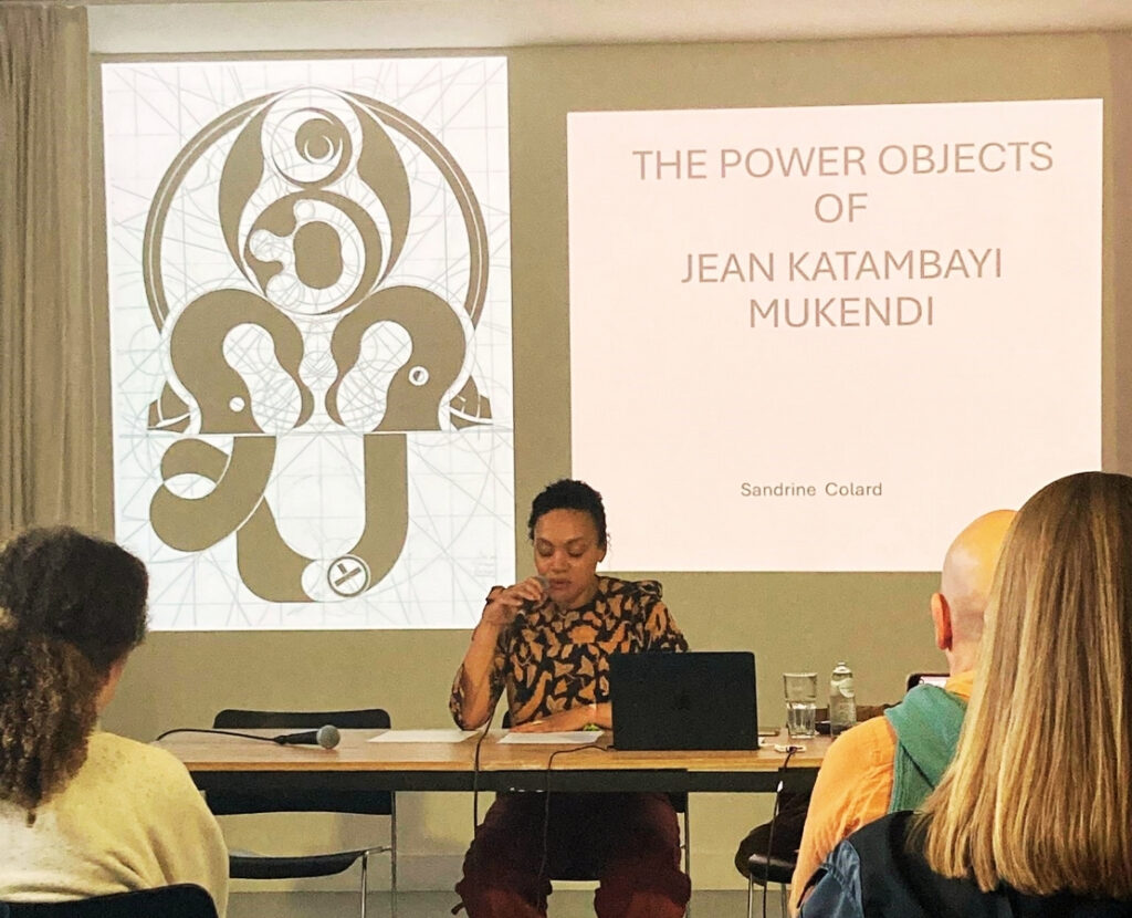 A woman sitting at a table giving a presentation to an audience, with a projected slide titled "THE POWER OBJECTS OF JEAN KATAMBAYI MUKENDI."