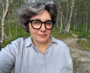 A person wearing dark-framed glasses and a light gray button-up shirt takes a selfie on a rocky path in a wooded area.